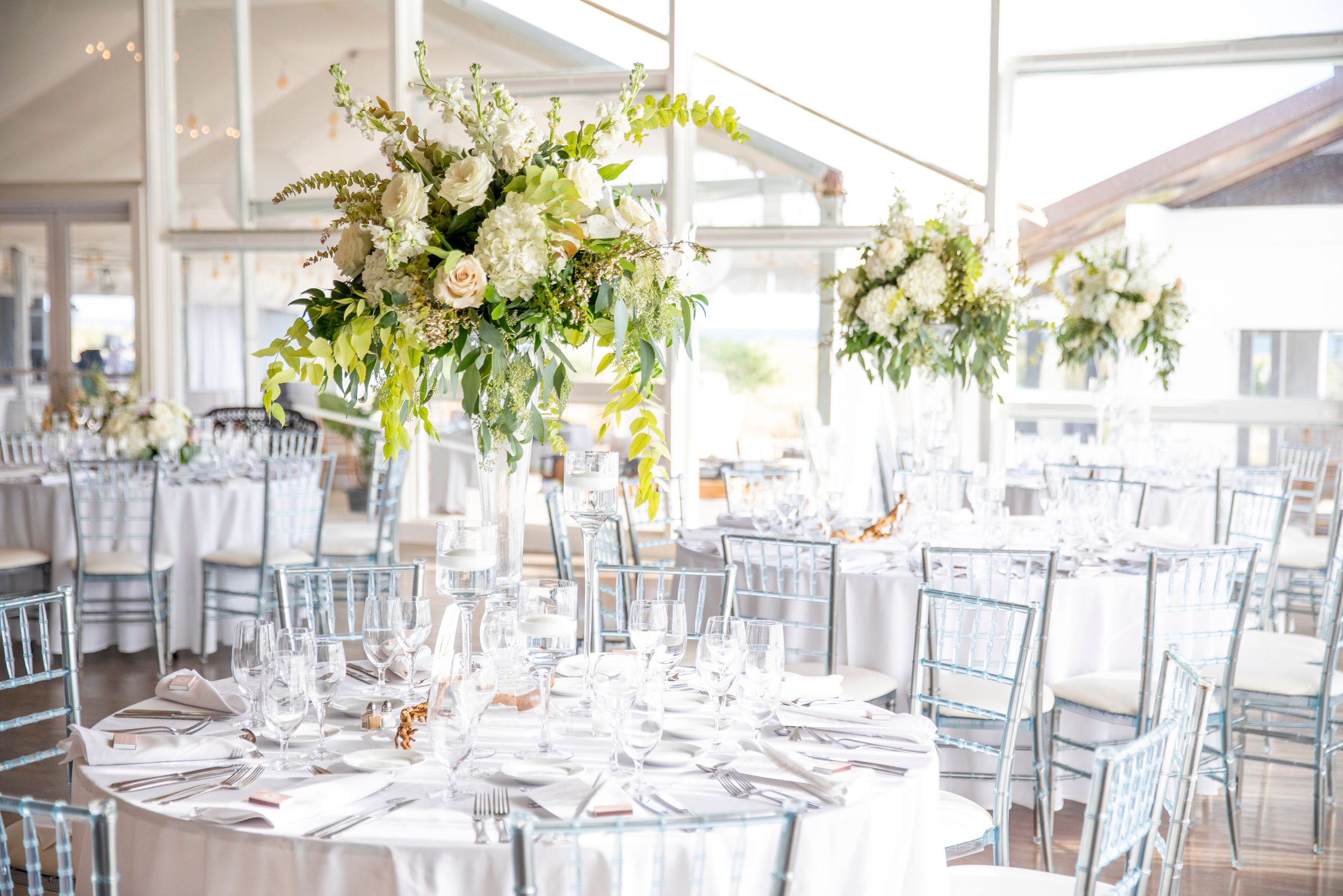 Elegant wedding reception setup with tall floral centerpieces and silver chiavari chairs.