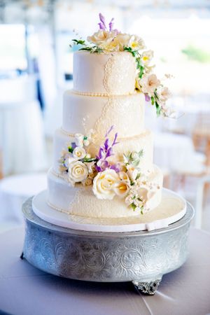 Four-tier white wedding cake decorated with fresh flowers in cream and purple tones.