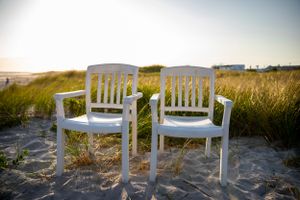 Two white wooden chairs sit empty on beach sand with beach grass in background.
