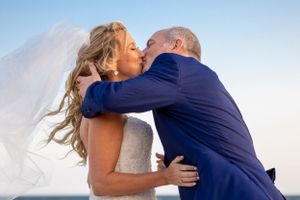 Romantic wedding kiss captured against coastal backdrop with bride's veil flowing in the breeze.