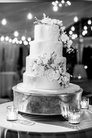 Elegant white three-tier wedding cake with cascading floral decorations displayed on silver stand with candlelit ambiance.