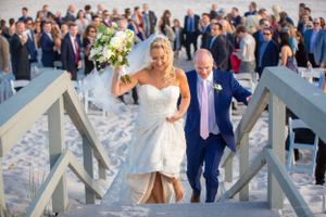 Happy couple descending white wooden stairs on a sunny beach with wedding guests in background.