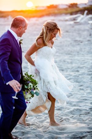 Couple walking barefoot on beach sand during golden hour sunset wedding celebration.