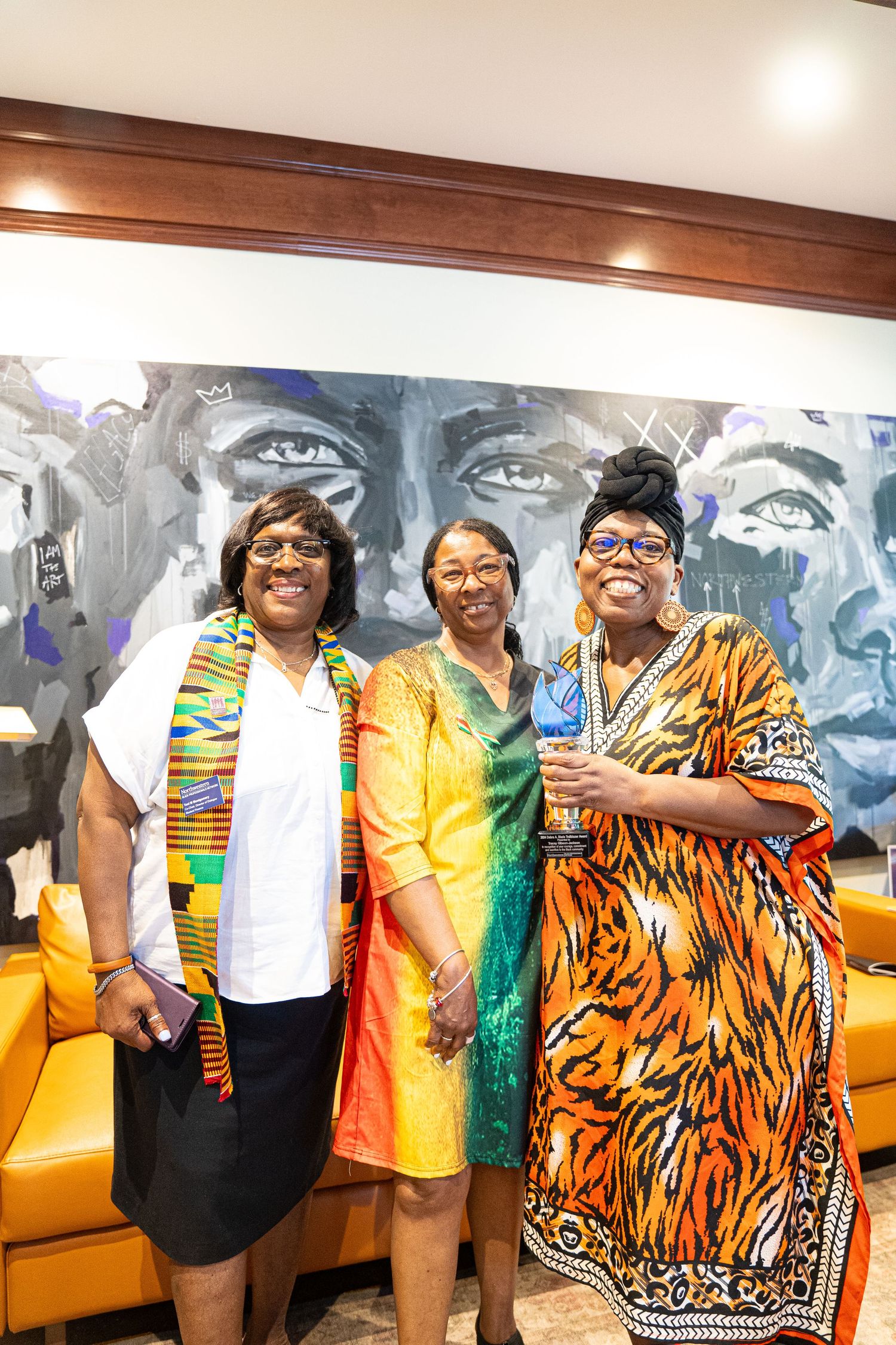 Three women in colorful traditional African-inspired clothing pose in front of a mural.