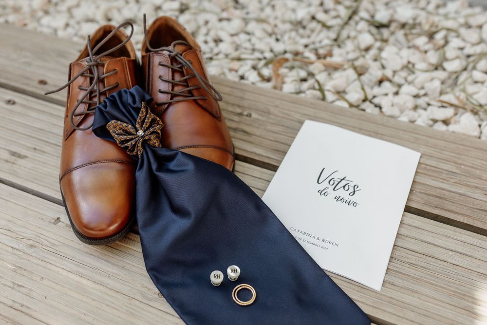 Brown leather dress shoes with cufflinks and a navy tie on a wooden surface next to a wedding invitation.