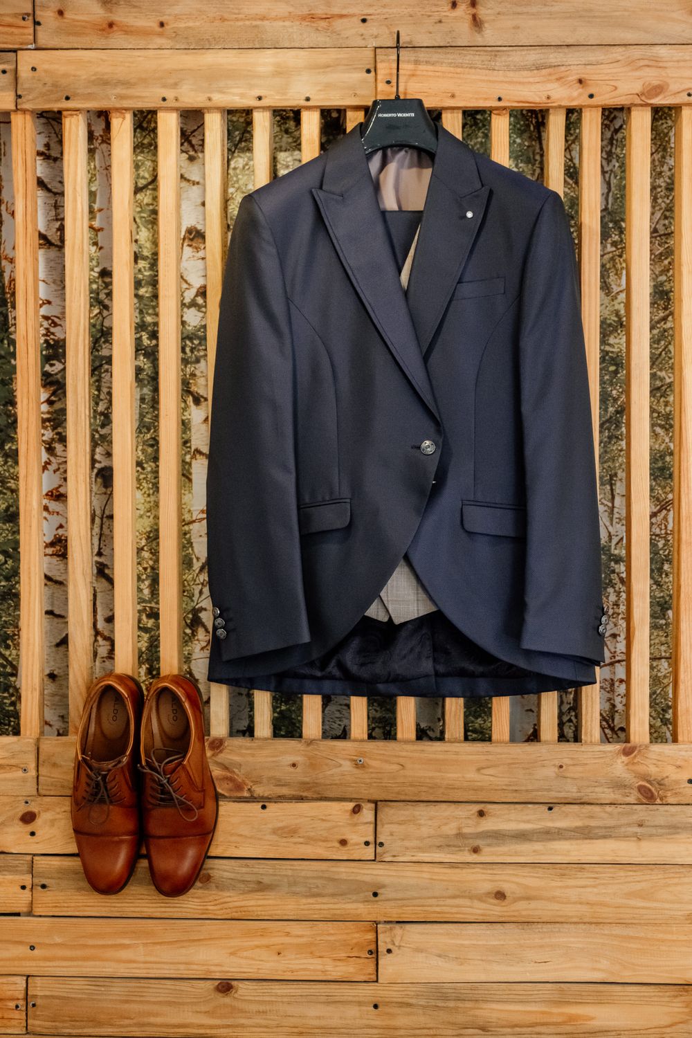 Navy suit jacket and brown leather shoes arranged on wooden backdrop.