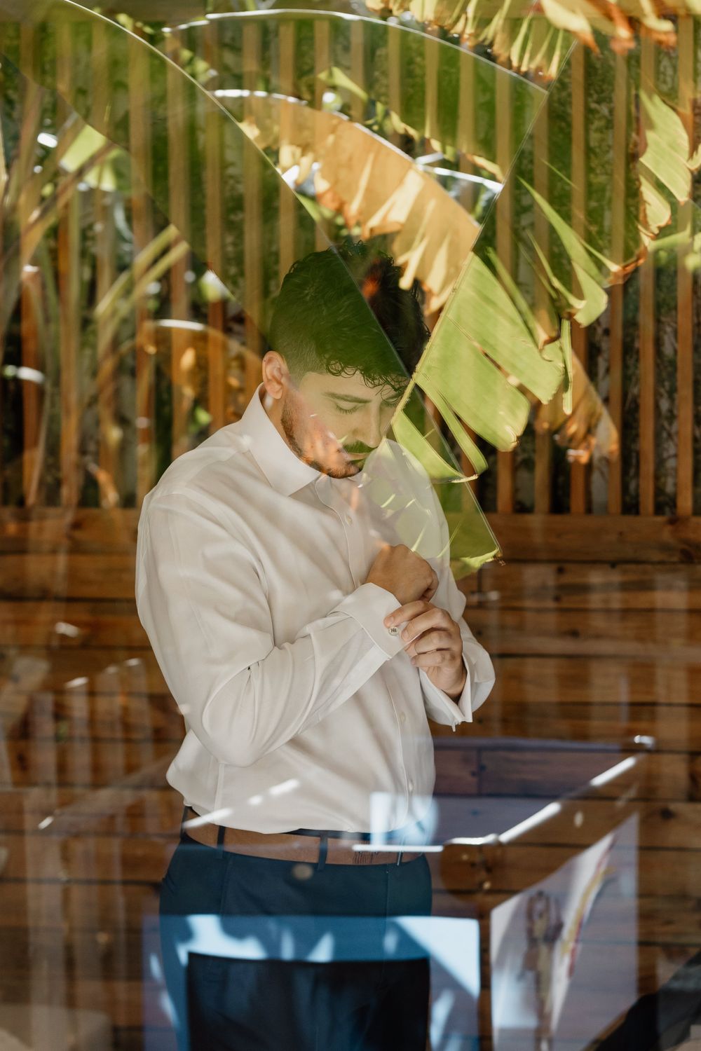 Person in white dress shirt buttoning up under tropical palm leaves.