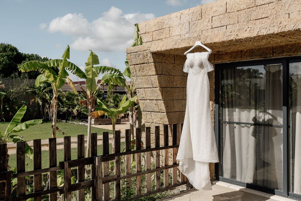 White wedding dress hanging outside a modern stone building with tropical plants.