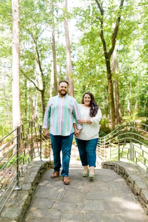 A couple walking hand in hand across a stone bridge surrounded by lush green trees.