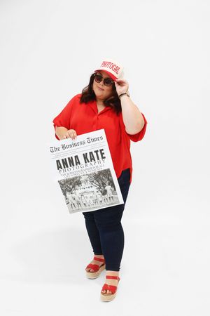 Woman in red shirt holding The Business Times newspaper featuring Anna Kate Photography, wearing sunglasses and red sandals.