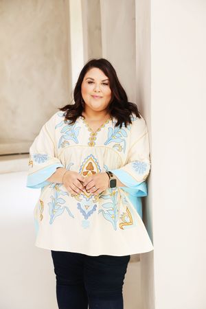 Woman in embroidered cream tunic top with blue floral design and dark jeans, smiling and leaning against a wall.