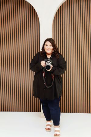 A smiling plus-size woman in black outfit holds a camera, standing before a modern wood slat wall with arch details.