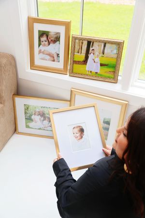 Woman in black jacket holding a gold-framed baby portrait surrounded by multiple framed children photos near a bright window.