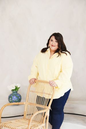 Plus-size woman in yellow shirt and jeans posing with rattan chair near blue vase on white background.