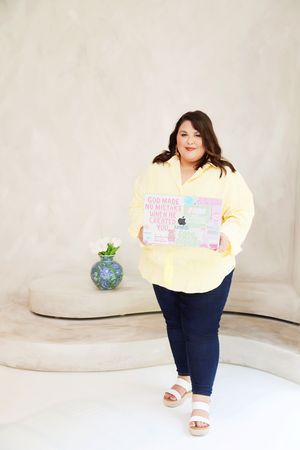 Plus-size woman in yellow shirt and navy jeans holds decorated laptop, standing in bright minimalist space.