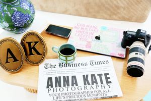 Anna Kate Photography branding flat lay with newspaper, monogram coasters, Canon camera, MacBook, and coffee on wooden desk.