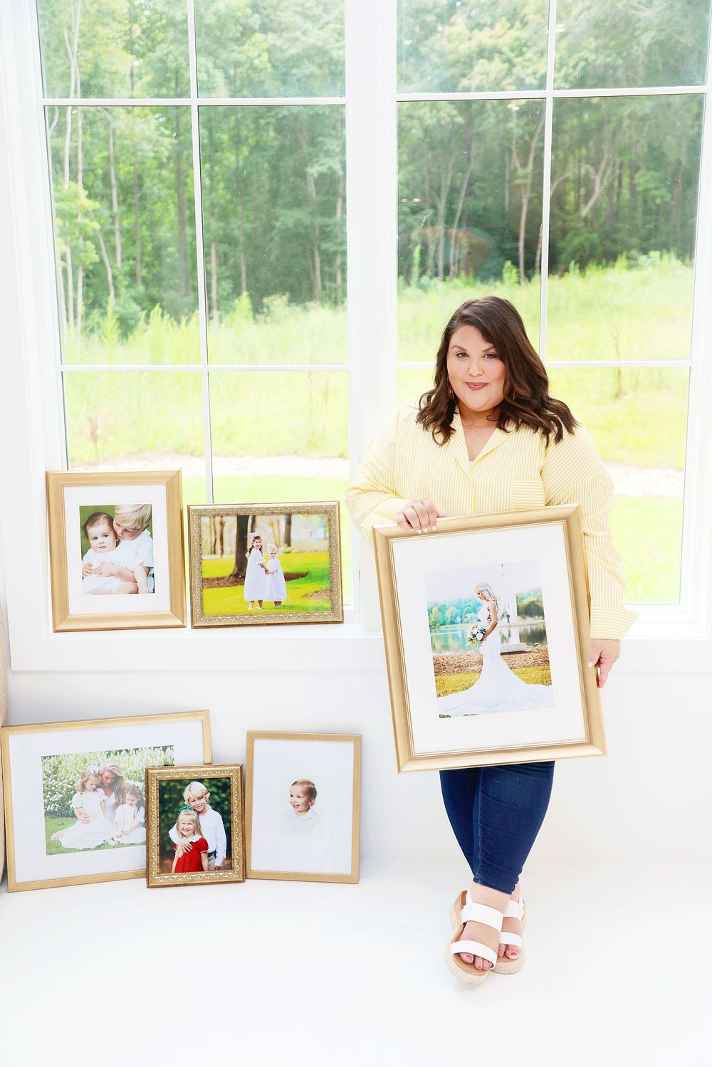 Woman in yellow blouse holding gold-framed family portrait surrounded by framed photos near bright window.
