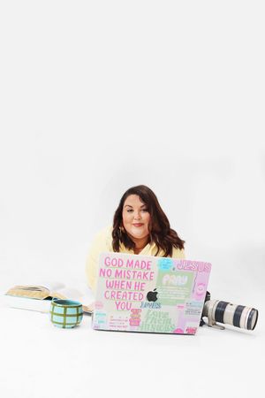 Woman sitting on floor with colorful sticker-covered laptop displaying faith-based messages, beside camera and mug.
