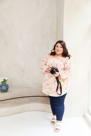 Woman in floral top and jeans holds camera, posing against neutral curved wall backdrop.