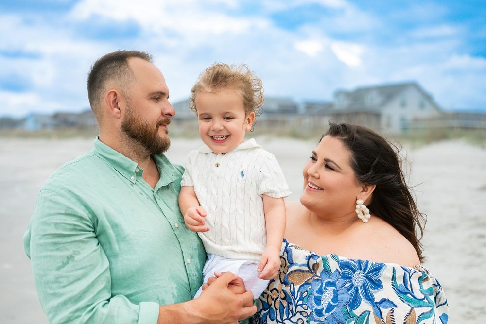 Happy family of three smiling on a beach, parents holding toddler, wearing colorful summer outfits with houses in background.