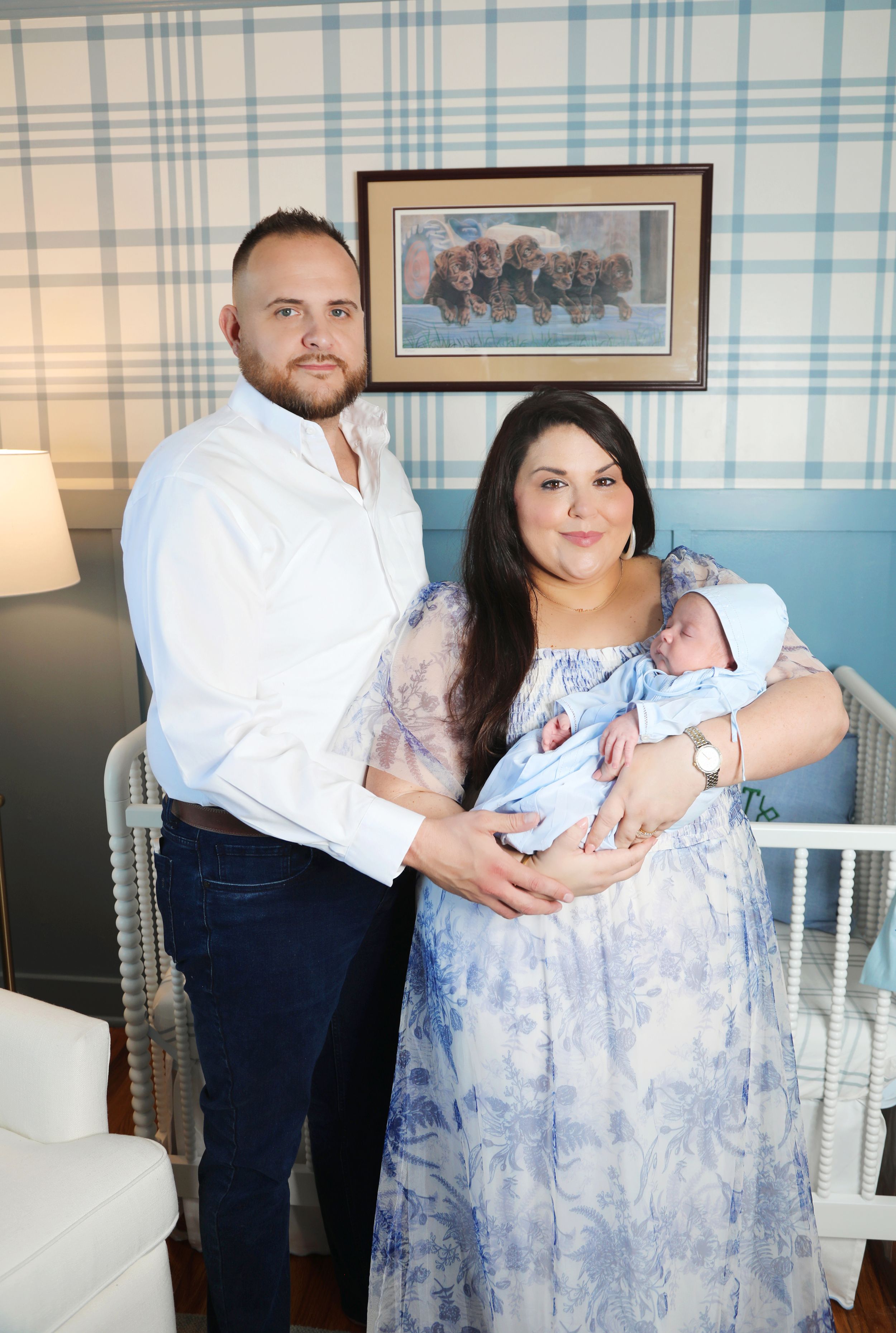A couple holds their newborn baby in a nursery setting with blue wallpaper and white furniture.