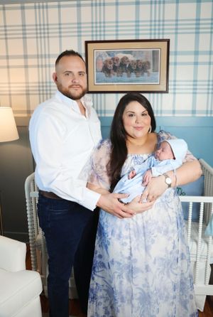 A couple holds their newborn baby in a nursery setting with blue wallpaper and white furniture.