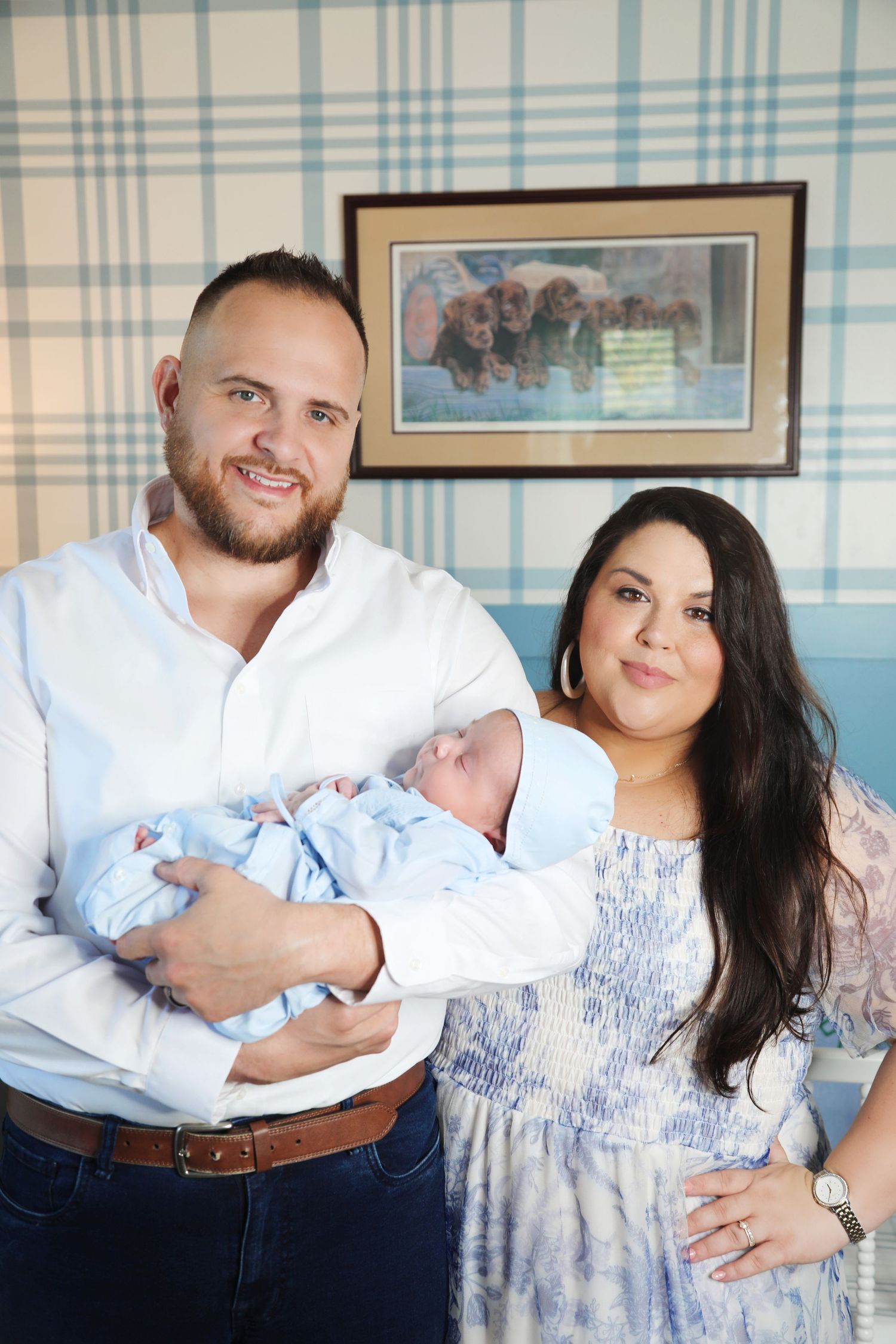 New parents with newborn baby posing against blue and white plaid wallpaper.