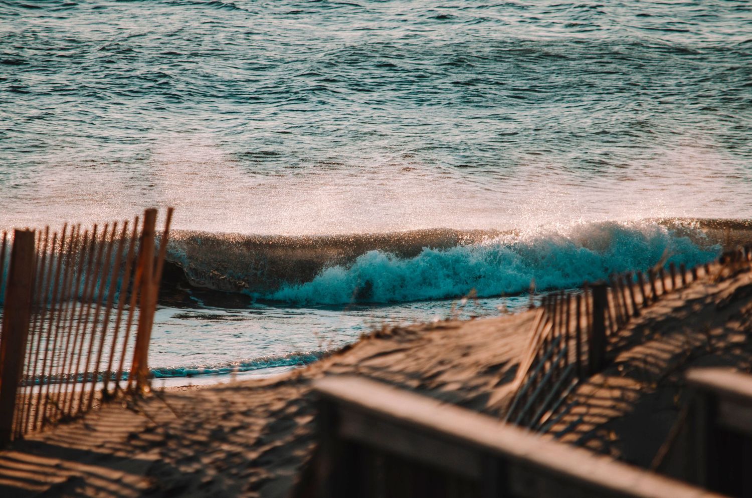 Waves crash along a sandy beach with wooden fencing during sunset.
