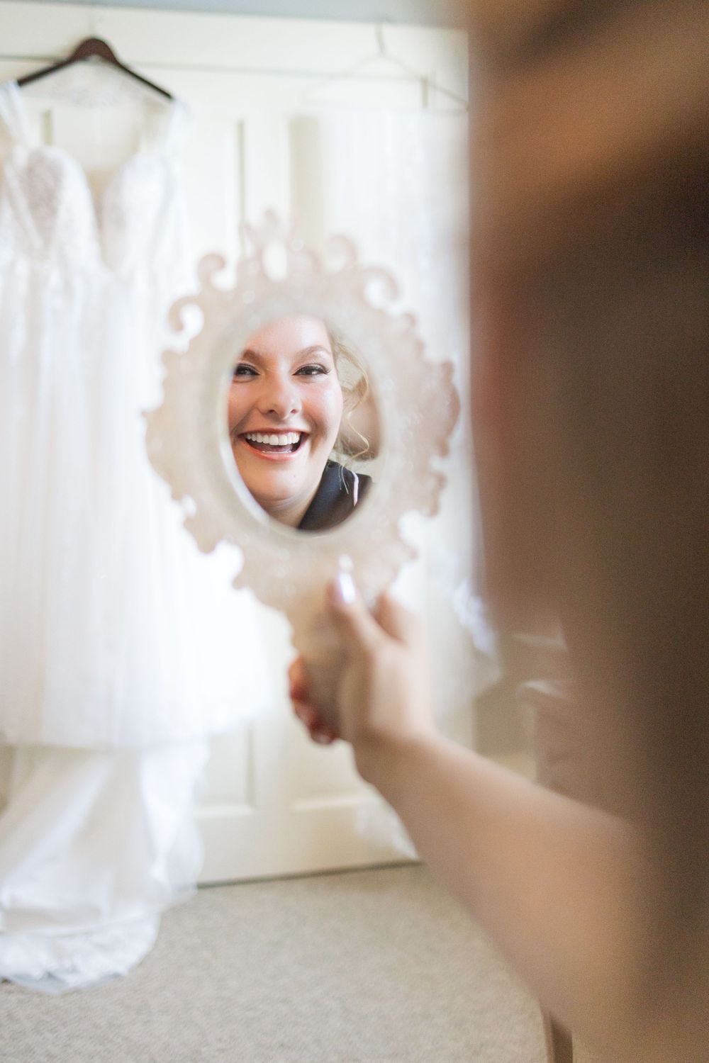 Reflection of joyful smile in ornate white handheld mirror with wedding dress in background.