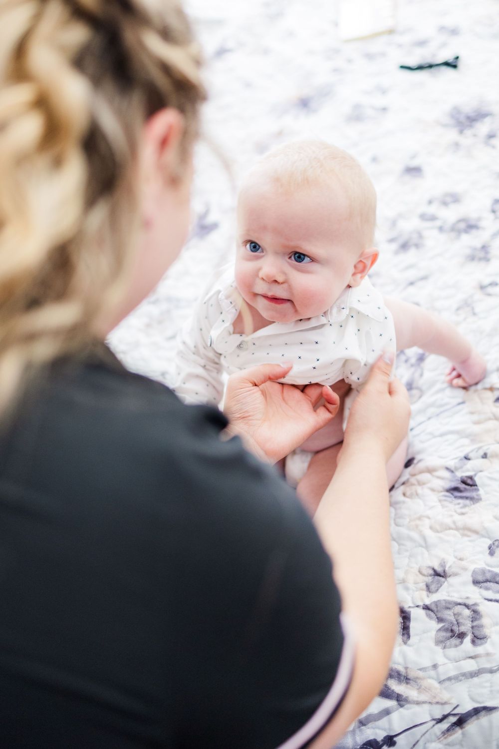 A smiling baby lying on a floral blanket being tickled by someone wearing black.
