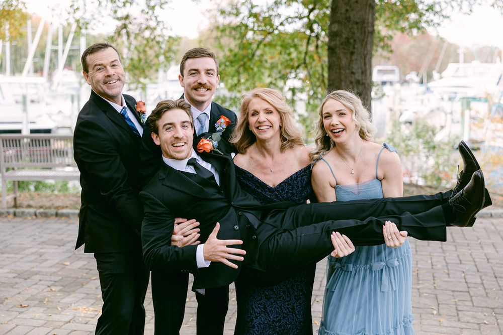 A group in formal attire poses playfully outdoors on a brick patio near trees and a marina.