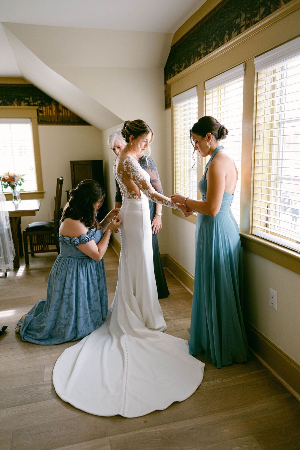 Bridesmaids in dusty blue dresses help the bride prepare in a sunlit room before the wedding ceremony.