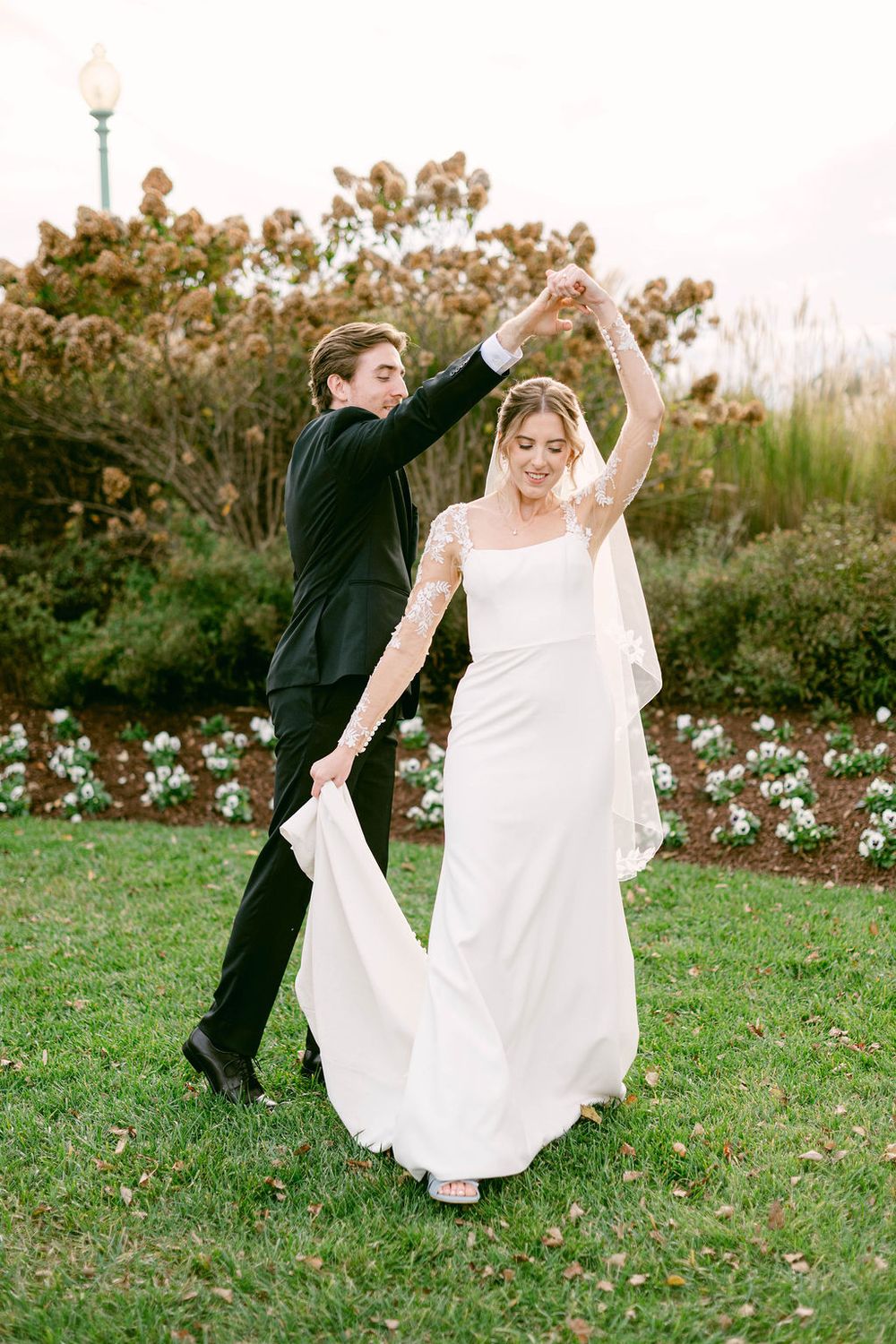Playful bride and groom dancing and twirling on grass in a garden setting.