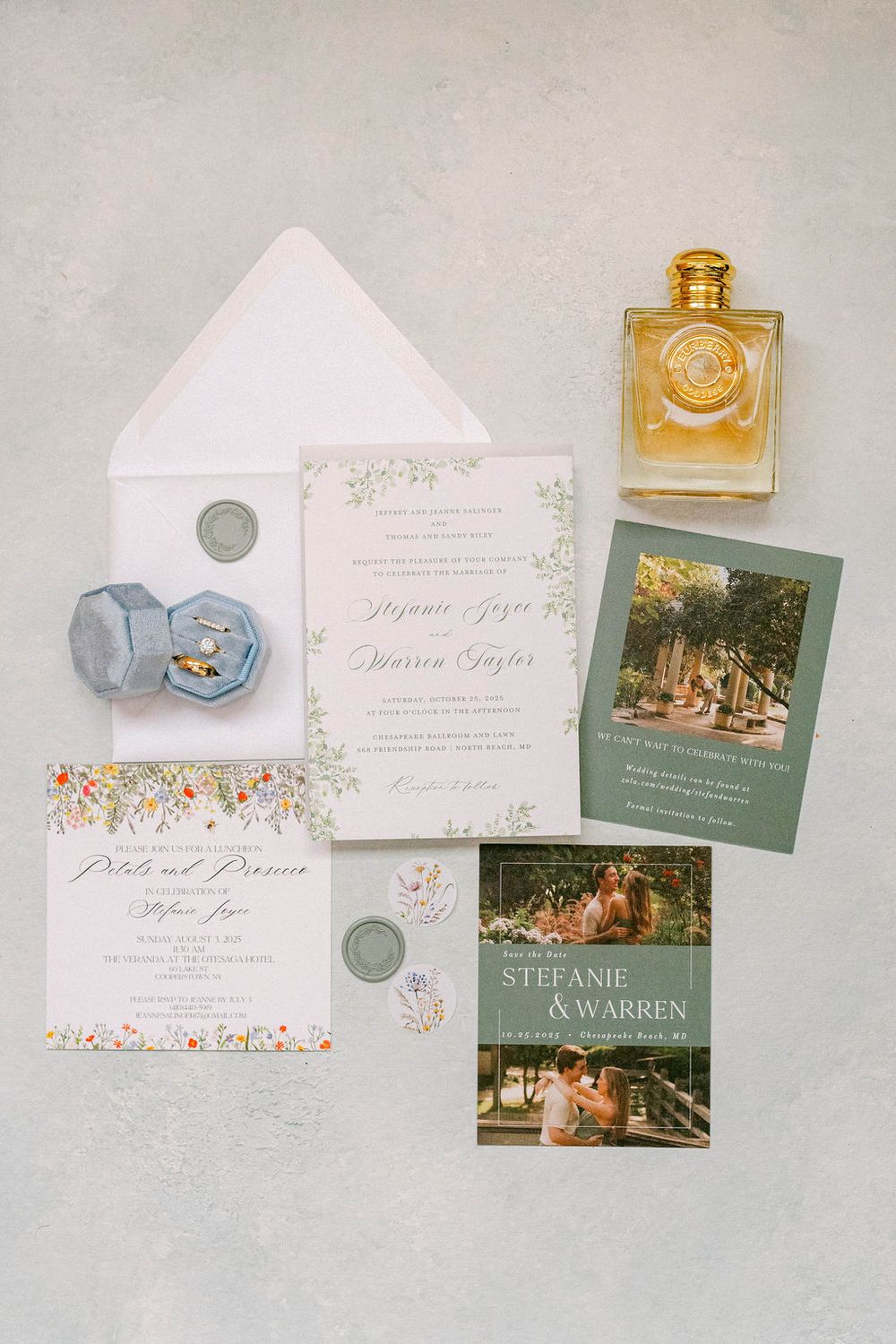 Wedding invitation suite with floral designs, photographs, and perfume bottles arranged on a light gray background.