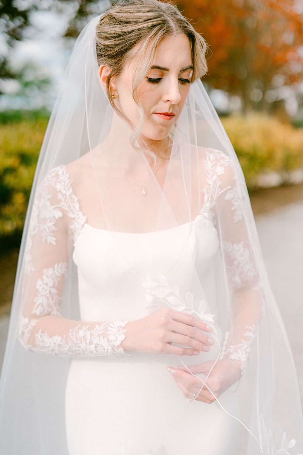 Elegant bride in white lace long sleeve wedding dress and sheer veil poses against autumn foliage background.