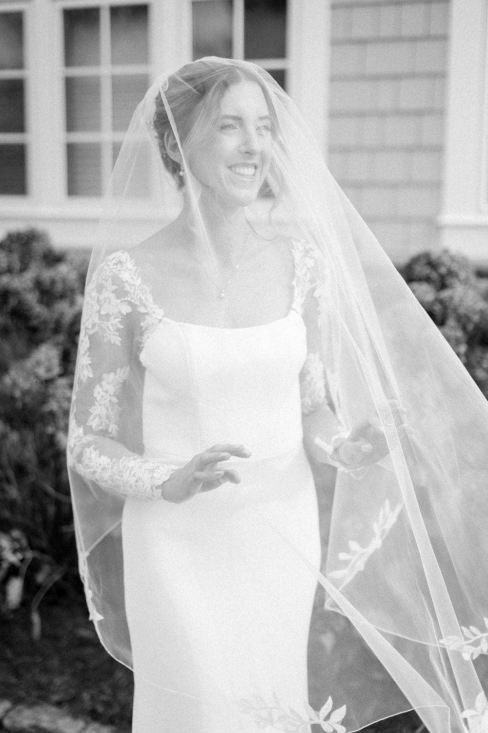 A bride in an elegant white wedding dress with lace sleeves and veil stands in front of a light-colored building.