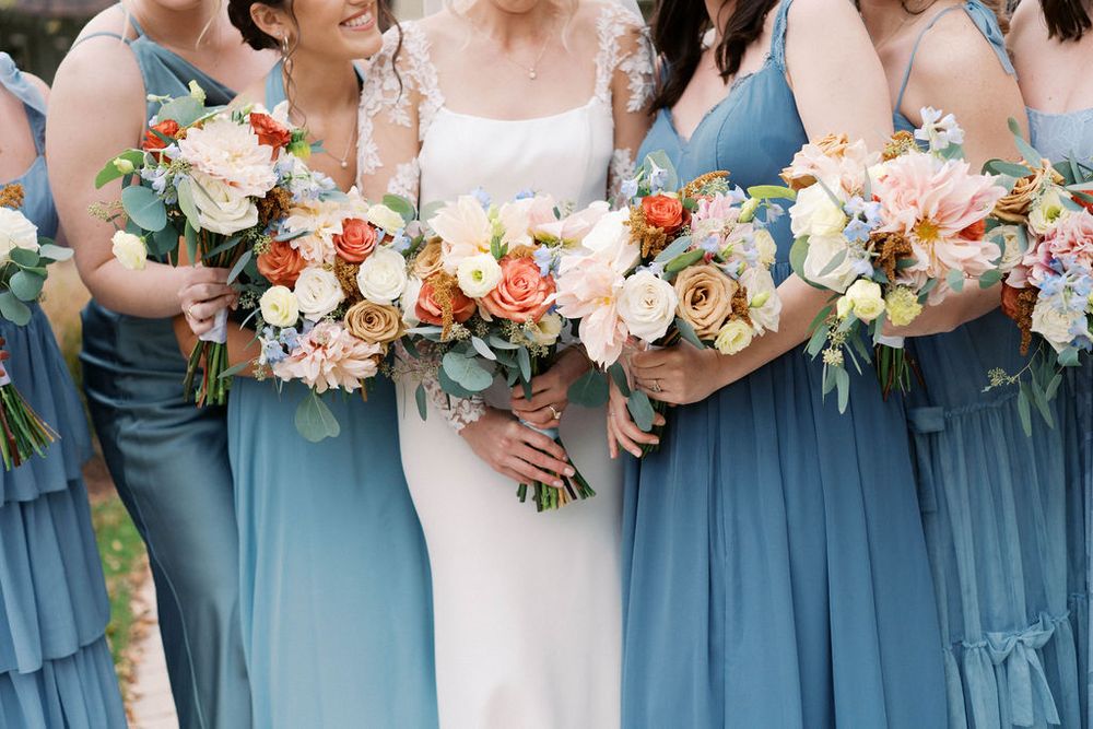 Bridesmaids in dusty blue dresses hold colorful peach and white floral bouquets at an outdoor wedding.