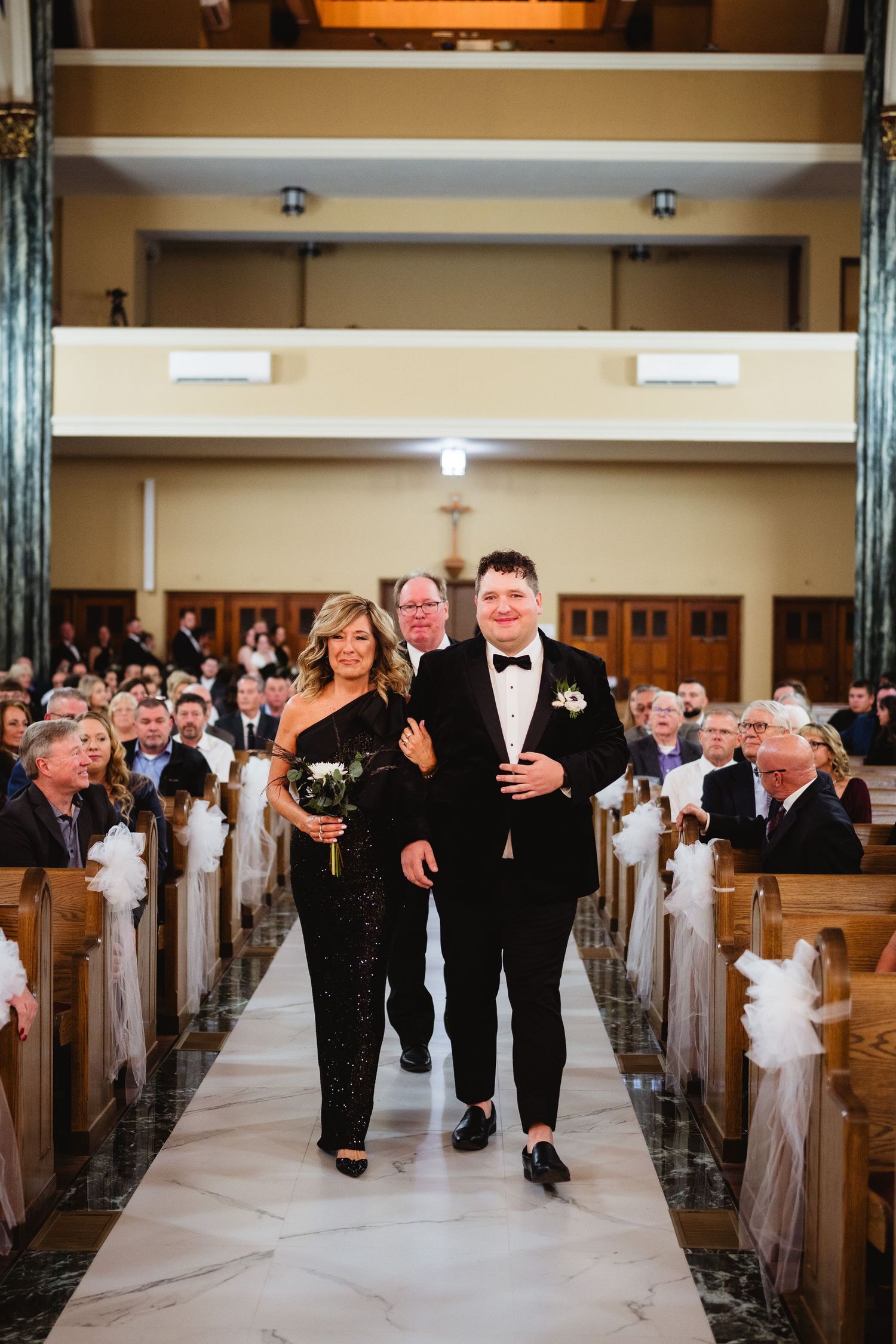 Wedding party members walk down the aisle of a traditional church with wooden pews decorated in white ribbons.