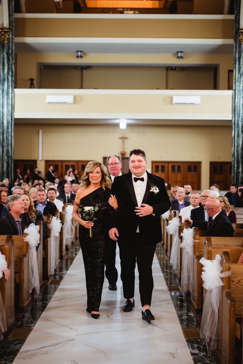 Wedding party members walk down the aisle of a traditional church with wooden pews decorated in white ribbons.