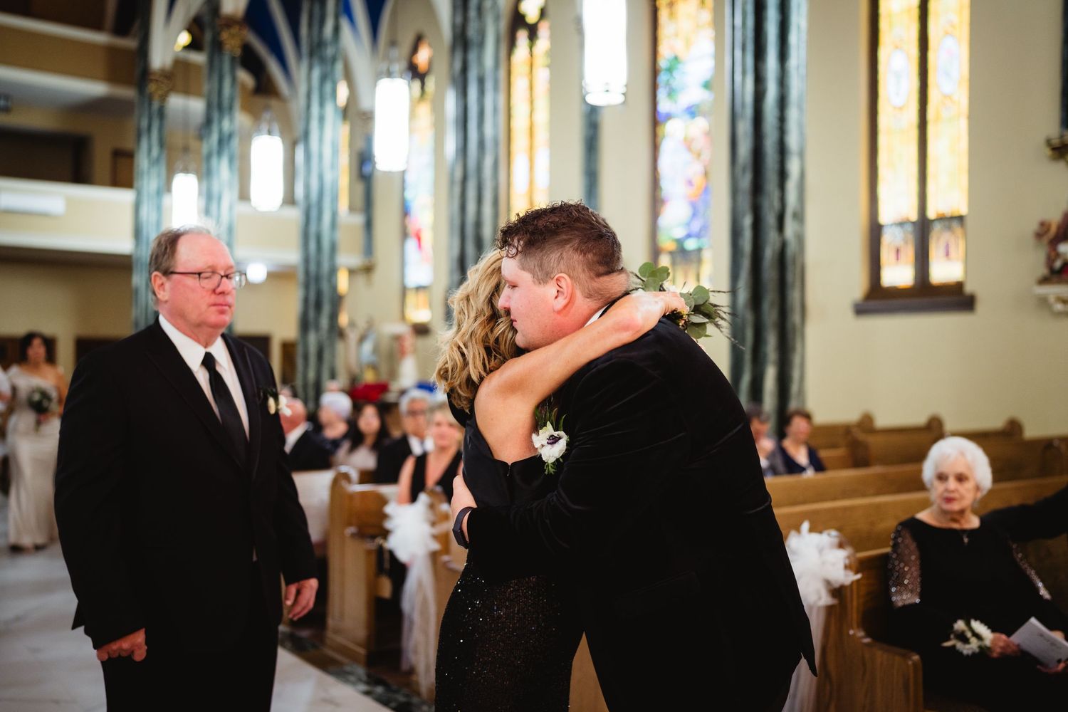 An emotional embrace takes place during a wedding ceremony inside a beautiful church with stained glass windows and gothic architecture.