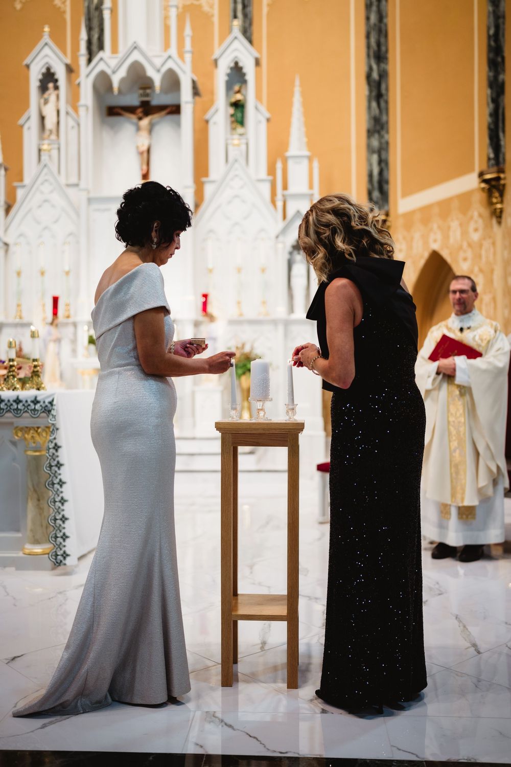 Two people stand at a wooden table during a Catholic church ceremony with a priest and crucifix visible in the background.