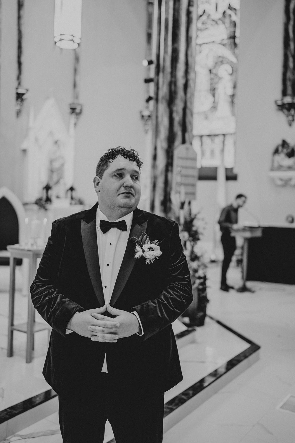 Man in black tuxedo and bow tie stands between white pillars in formal setting.