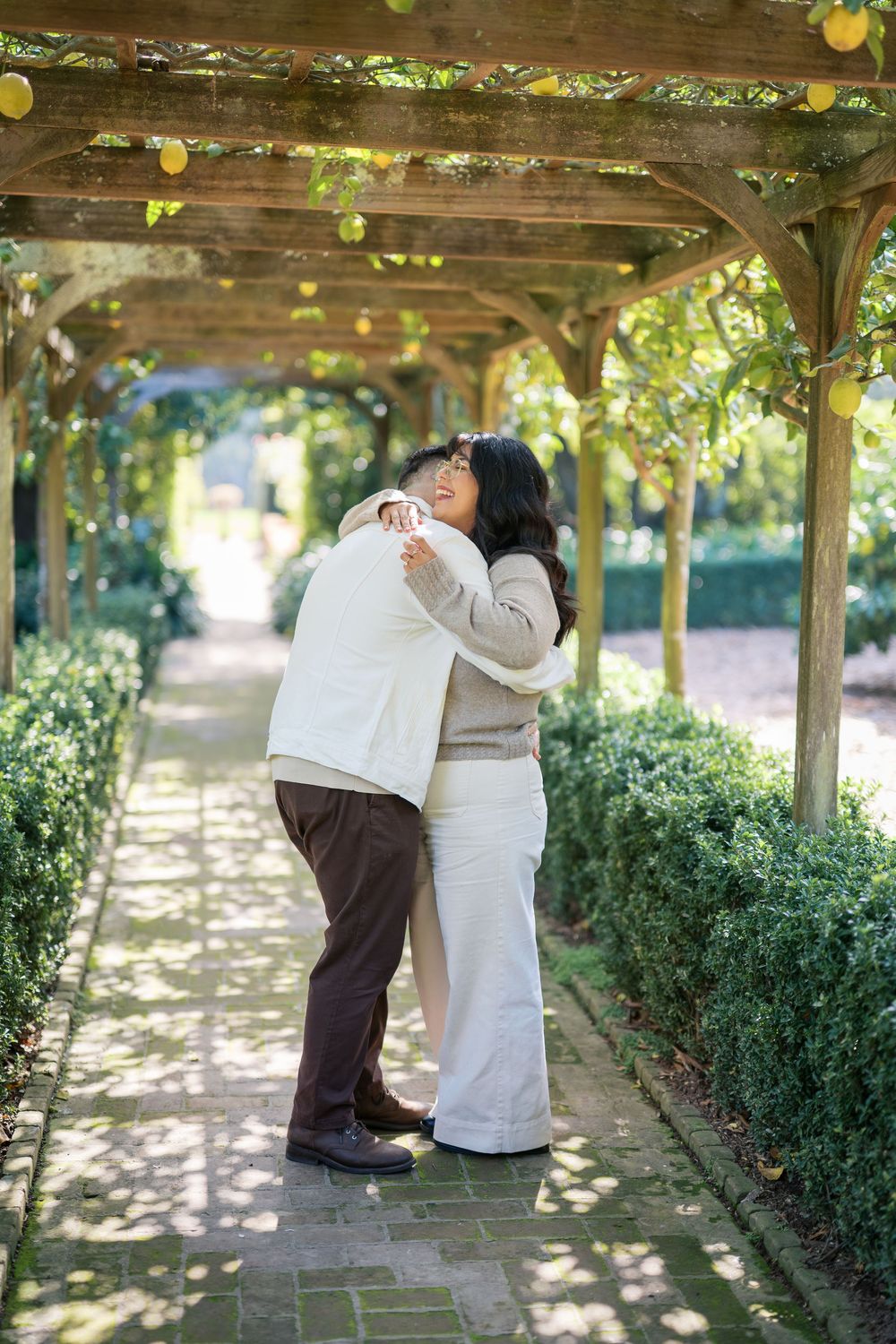 A romantic couple embraces under a garden pergola covered in vines and greenery on a sunny afternoon.
