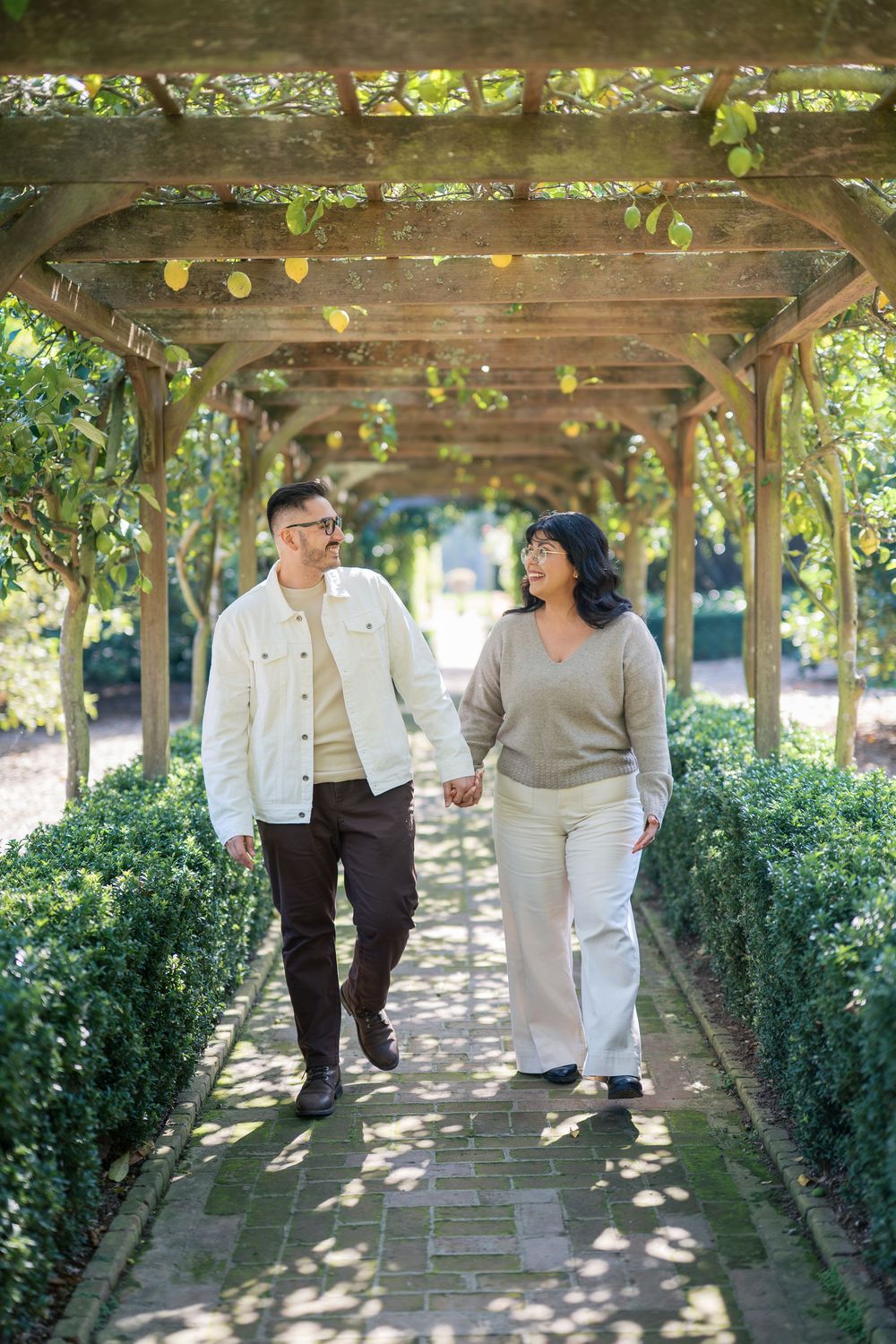 A couple walks together holding hands under a wooden pergola covered in string lights and greenery in a garden setting.