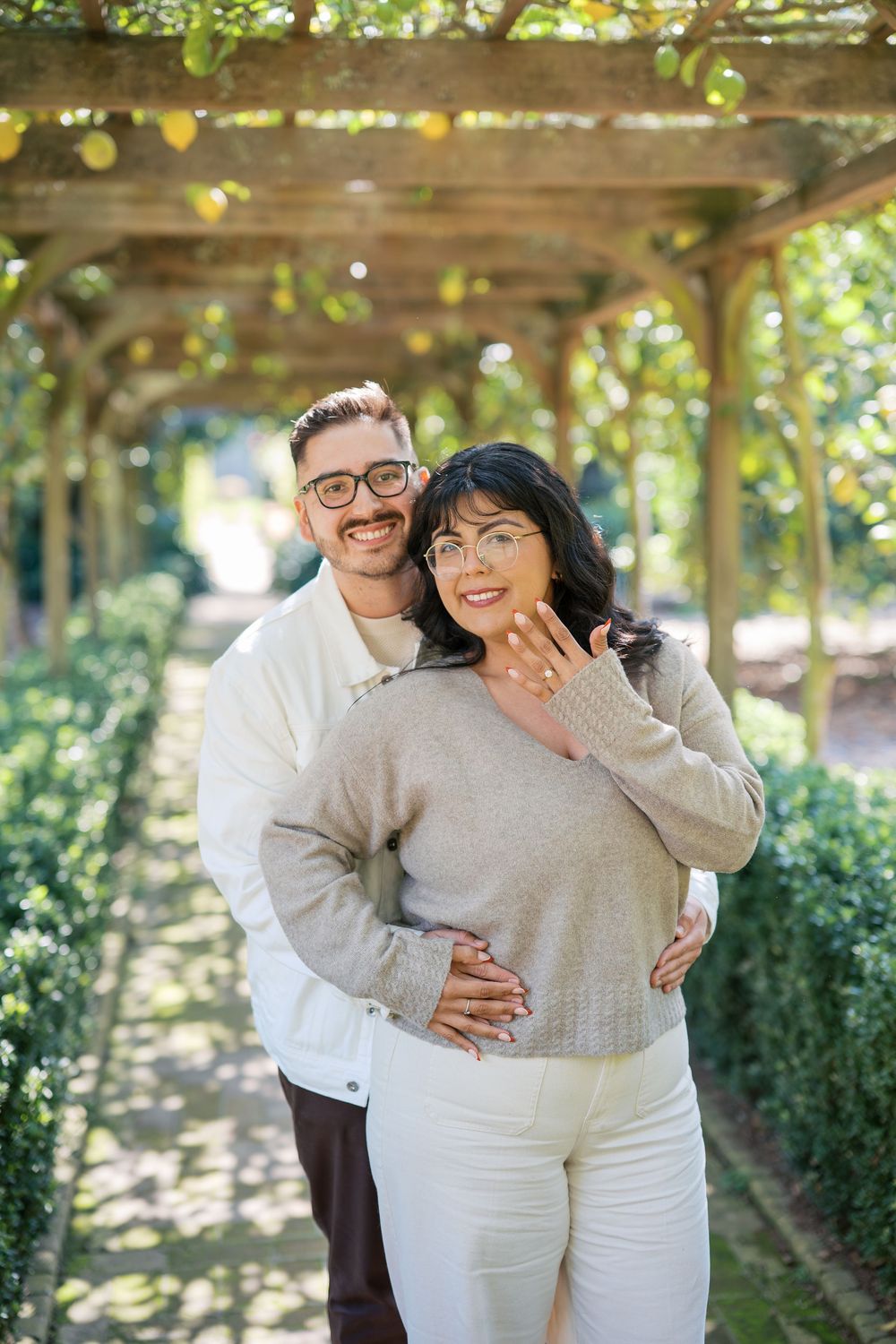 A couple wearing matching white and gray outfits embrace and laugh together under a pergola in a garden setting.