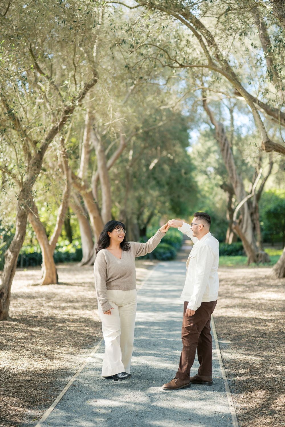 A couple dancing playfully on a tree-lined path in a park on a sunny day.