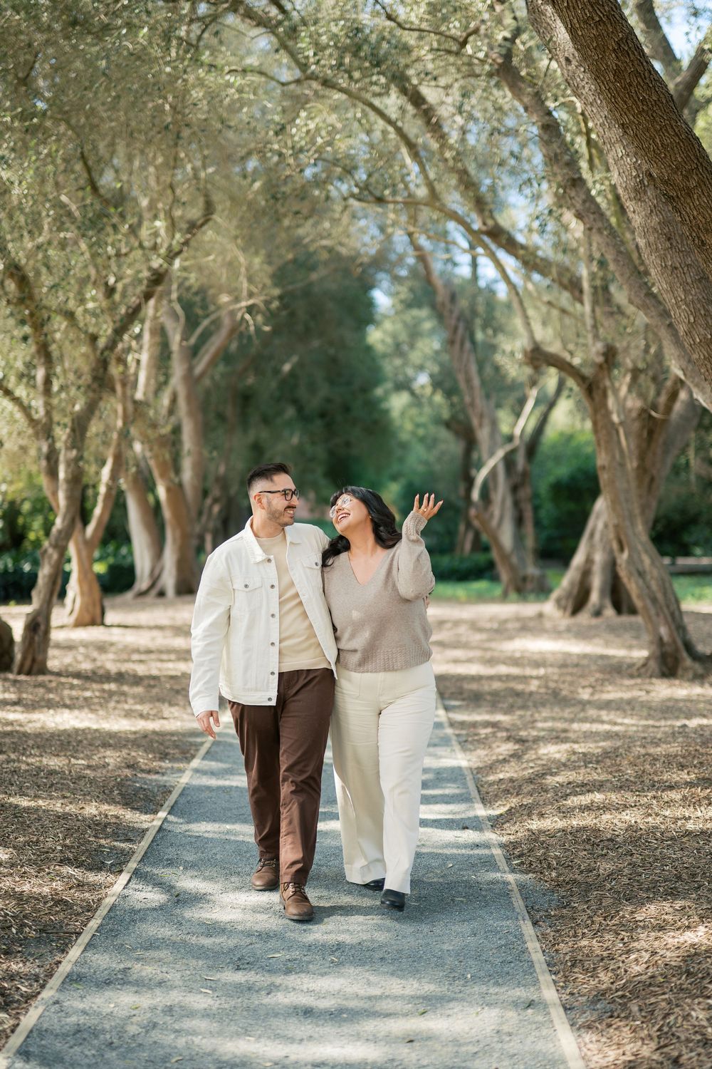 A couple walks and laughs together on a tree-lined path in a romantic outdoor setting.