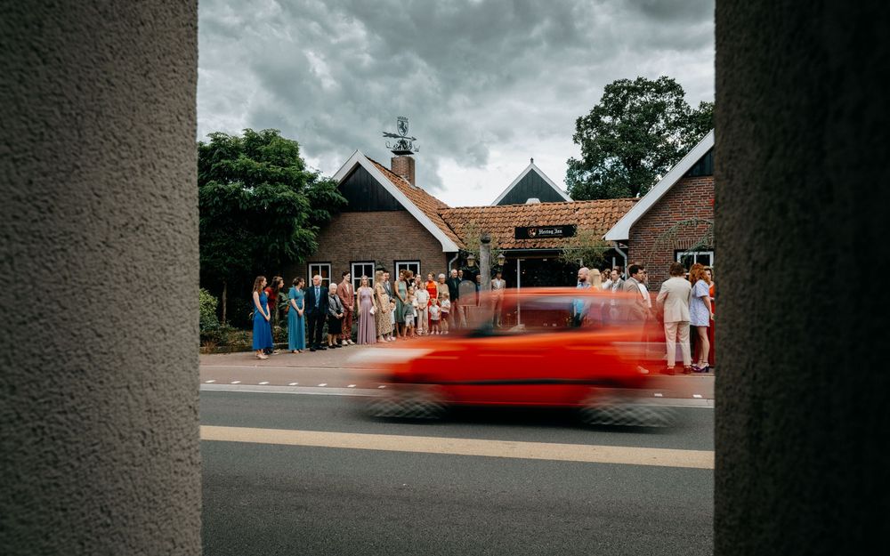 De gasten voor de bruiloft wachten de aankomst van het bruidspaar af terwijl een auto voorbij raast.