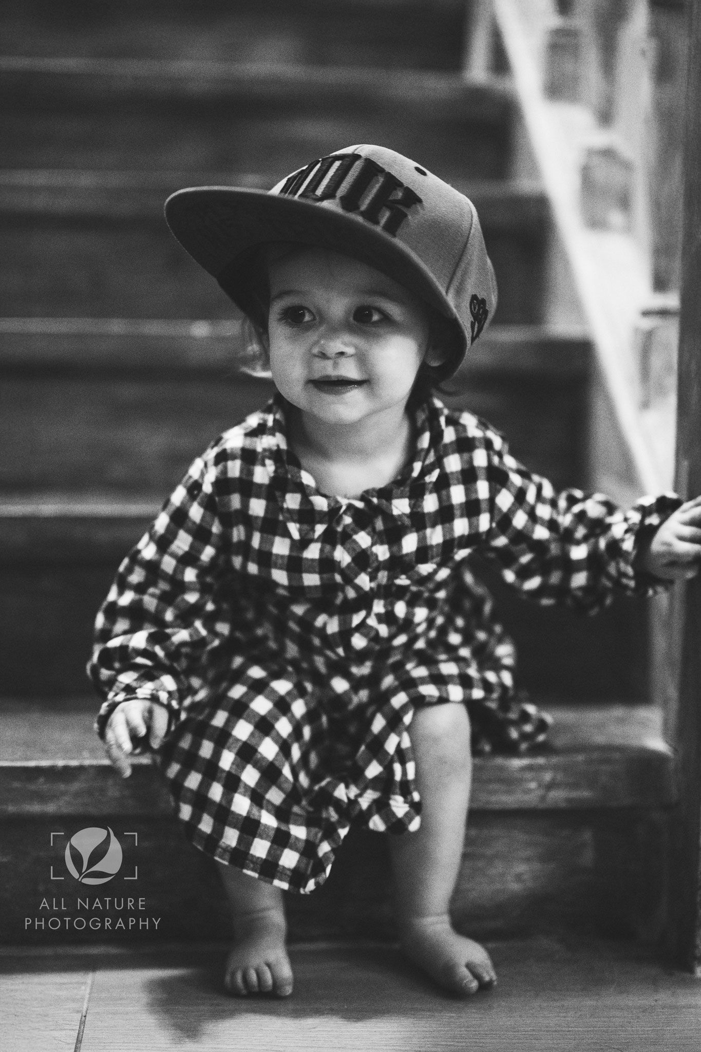 A black and white photo of a small child in a plaid dress and baseball cap sitting on wooden stairs.