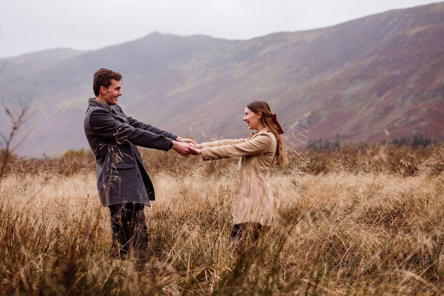 A couple holds hands while standing in a Lake District field of tall grass with misty mountains in the background.